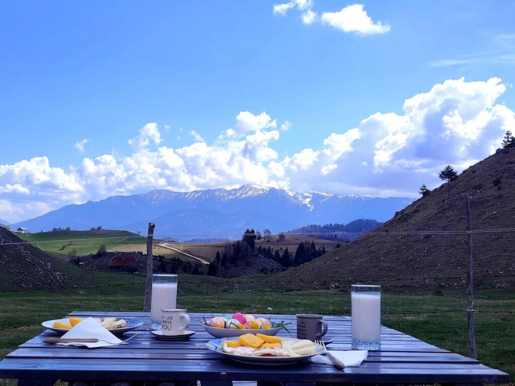 Masă de mic dejun în aer liber, cu produse tradiționale și fundal montan spectaculos, fotografiată la Cabana Alloria.